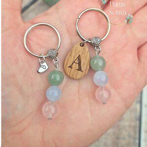 May include: Two keychains with wooden charms engraved with the letters A and B. Each charm is attached to a silver ring with a beaded chain of green, blue, and pink stones.