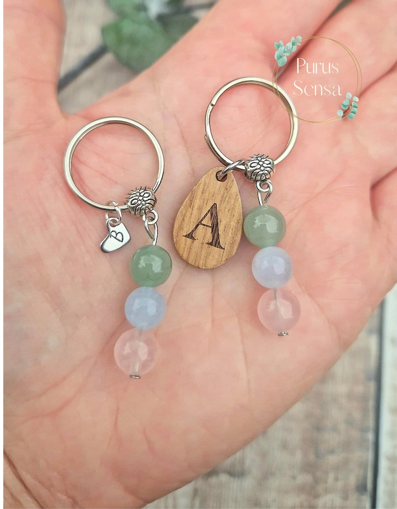 May include: Two keychains with wooden charms engraved with the letters A and B. Each charm is attached to a silver ring with a beaded chain of green, blue, and pink stones.