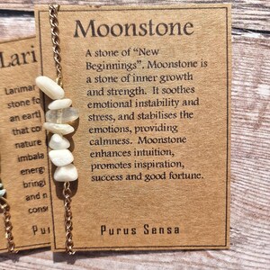 May include: A brown card with the text "Moonstone" in large white letters. The card describes moonstone as a stone of "New Beginnings" and lists its properties. The card also includes a gold chain with white moonstone beads.