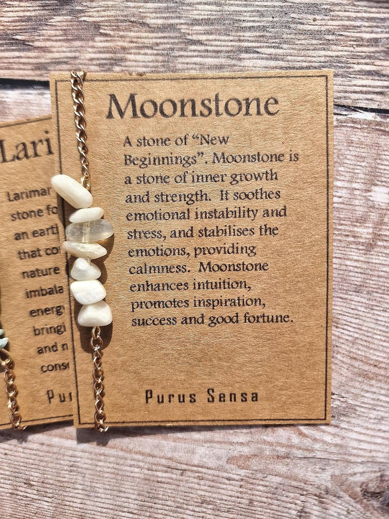 May include: A brown card with the text "Moonstone" in large white letters. The card describes moonstone as a stone of "New Beginnings" and lists its properties. The card also includes a gold chain with white moonstone beads.
