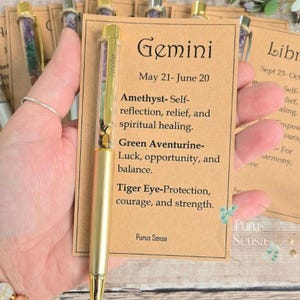 Könnte beinhalten: Ein goldfarbener Stift mit einem transparenten Schaft, gefüllt mit bunten Steinen, neben einer Karte mit dem Wort "Gemini" und Text. Der Stift trägt den Aufdruck "Jennifer".