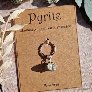 May include: Pyrite keychain with a silver ring and a small, rough, green and gold stone. The card reads "Pyrite Abundance. Confidence. Protection. Purus Sensa."