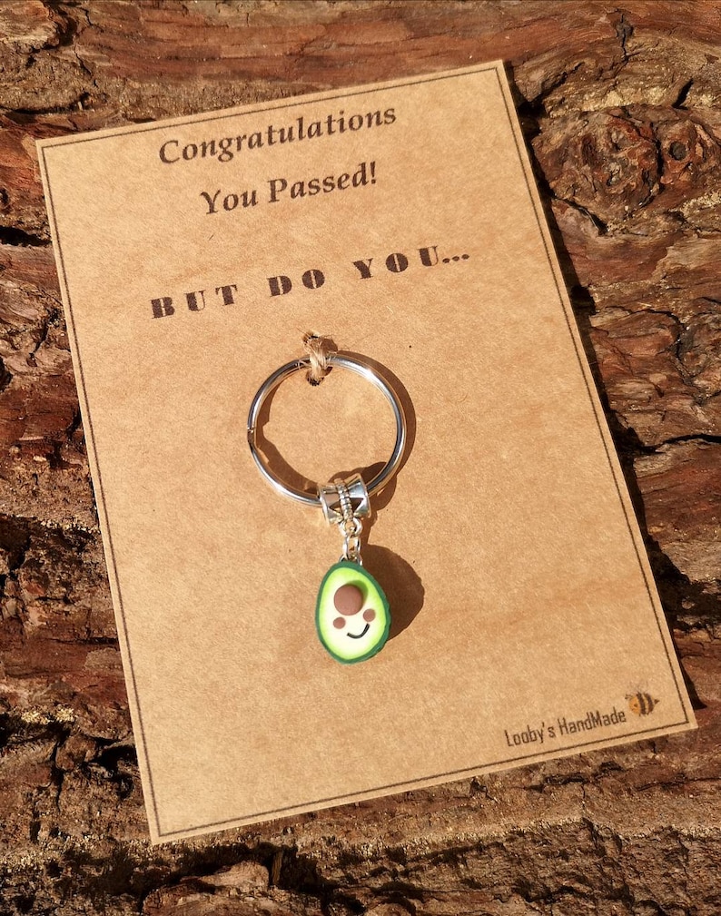 May include: A brown card with the text "Congratulations You Passed! BUT DO YOU..." and a silver key chain with a green avocado charm with a brown pit and a smiling face. The card has the text "Loopy's HandMade" in the bottom right corner.