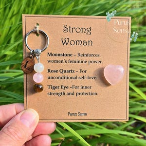 May include: A keychain with a wooden letter "L" charm, moonstone, rose quartz, and tiger eye beads. The card reads "Strong Woman" with descriptions of each stone's properties. A small pink heart-shaped stone is also included.
