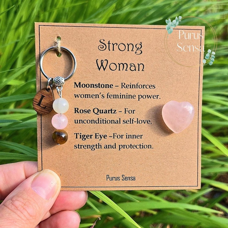 May include: A keychain with a wooden letter "L" charm, moonstone, rose quartz, and tiger eye beads. The card reads "Strong Woman" with descriptions of each stone's properties. A small pink heart-shaped stone is also included.