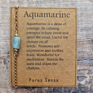 May include: A gold chain necklace with a light blue aquamarine stone pendant. The card attached to the necklace reads "Aquamarine is a stone of courage. Its calming energies reduce stress and quiet the mind. Useful for closure on all levels. Promotes self-expression and soothes fears. Wonderful for meditation. Shields the aura and aligns the chakras. Purus Sensa."