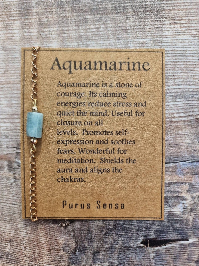May include: A gold chain necklace with a light blue aquamarine stone pendant. The card attached to the necklace reads "Aquamarine is a stone of courage. Its calming energies reduce stress and quiet the mind. Useful for closure on all levels. Promotes self-expression and soothes fears. Wonderful for meditation. Shields the aura and aligns the chakras. Purus Sensa."