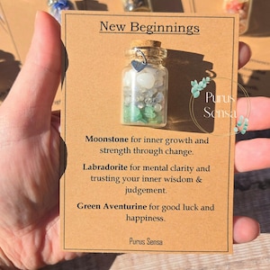 May include: A small glass bottle filled with various crystals and topped with a cork stopper. The bottle is labeled "New Beginnings" and includes a description of the crystals: Moonstone for inner growth and strength through change, Labradorite for mental clarity and trusting your inner wisdom & judgement, Green Aventurine for good luck and happiness. The label also includes the text "Purus Sensa".