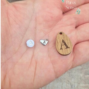 May include: Three charms with the letter A on them. One charm is a white round bead, one is a silver heart, and one is a wooden oval.