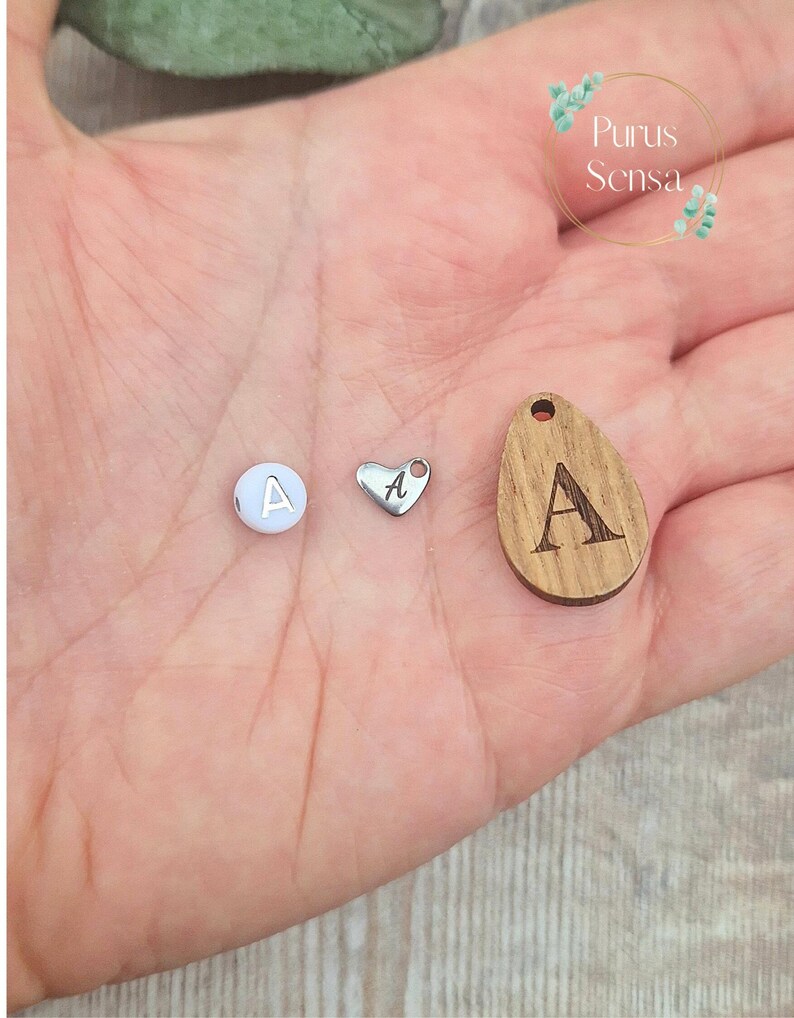 May include: Three charms with the letter A on them. One charm is a white round bead, one is a silver heart, and one is a wooden oval.