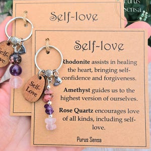 May include: A self-love keychain with a silver ring, a heart charm, and a wooden tag that says "Self-Love". The keychain also features a variety of crystals, including amethyst, rose quartz, and rhodonite. The card attached to the keychain reads "Self-love" and includes descriptions of the crystals and their meanings.