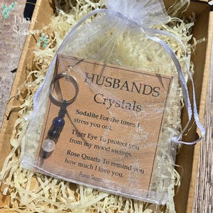 May include: A keychain with three crystals, including sodalite, tiger eye, and rose quartz, is displayed in a sheer white bag. The bag contains a card that reads "Husbands Crystals" with descriptions of each crystal's purpose.