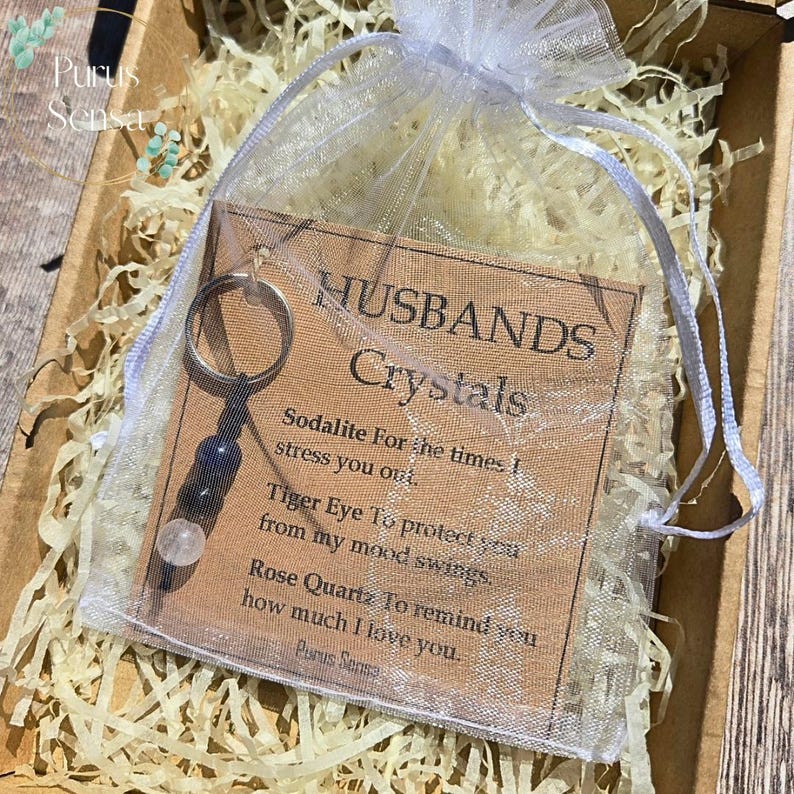 May include: A keychain with three crystals, including sodalite, tiger eye, and rose quartz, is displayed in a sheer white bag. The bag contains a card that reads "Husbands Crystals" with descriptions of each crystal's purpose.
