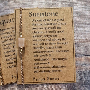 May include: A gold chain necklace with a sunstone pendant. The pendant is a light peach color. The card attached to the necklace reads "Sunstone: A stone of luck & good fortune. Sunstone clears and energises all the chakras. It instils good nature, heightens intuition and allows the real self to shine through happily. It acts as an antidepressant. Increases self-worth & confidence. Encourages optimism & enthusiasm. Stimulates self-healing powers. Purus Sensa."