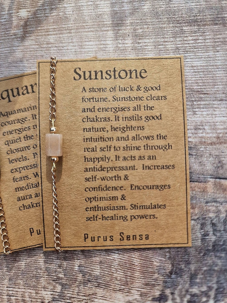 May include: A gold chain necklace with a sunstone pendant. The pendant is a light peach color. The card attached to the necklace reads "Sunstone: A stone of luck & good fortune. Sunstone clears and energises all the chakras. It instils good nature, heightens intuition and allows the real self to shine through happily. It acts as an antidepressant. Increases self-worth & confidence. Encourages optimism & enthusiasm. Stimulates self-healing powers. Purus Sensa."
