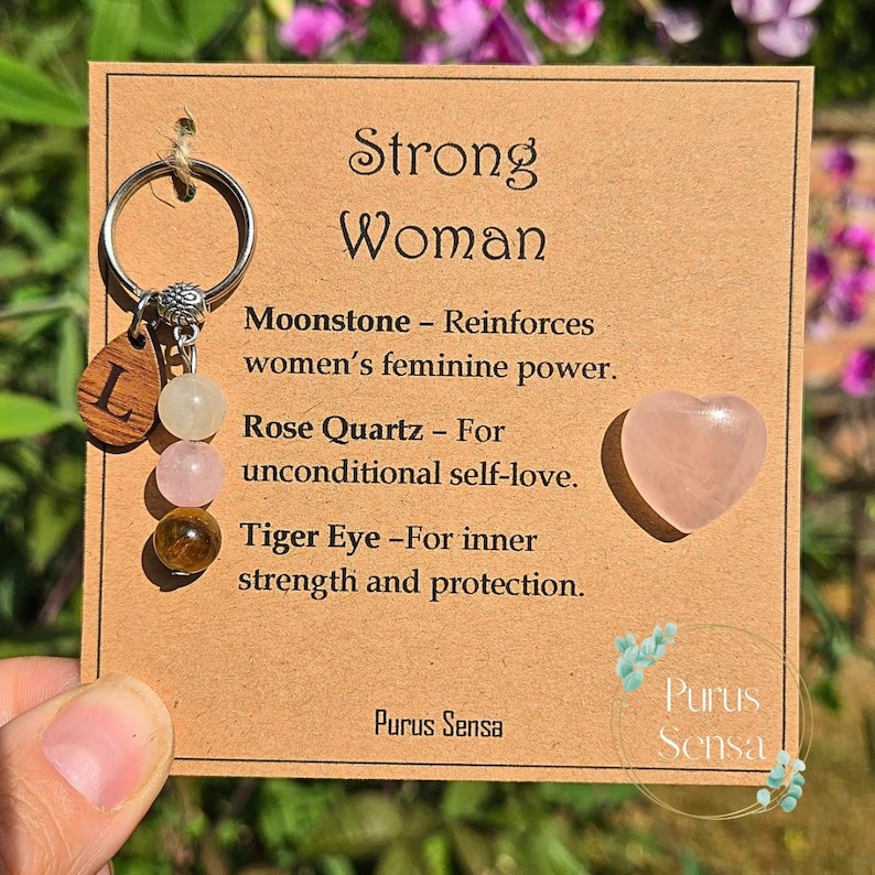 May include: A keychain with a wooden letter "L" charm, moonstone, rose quartz, and tiger eye beads. The card reads "Strong Woman" and lists the properties of each stone. A heart-shaped rose quartz is also included.
