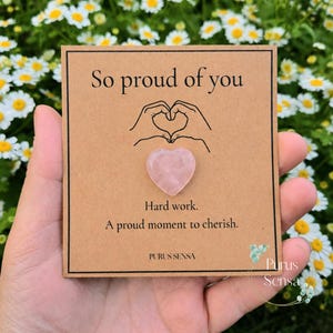 Könnte beinhalten: Eine quadratische, hellbraune Karte mit dem Text So proud of you, einer Strichzeichnung von Händen, die ein Herz formen, und einem kleinen, polierten, herzförmigen rosa Stein. Darunter steht Hard work. A proud moment to cherish. Purus Sensa.