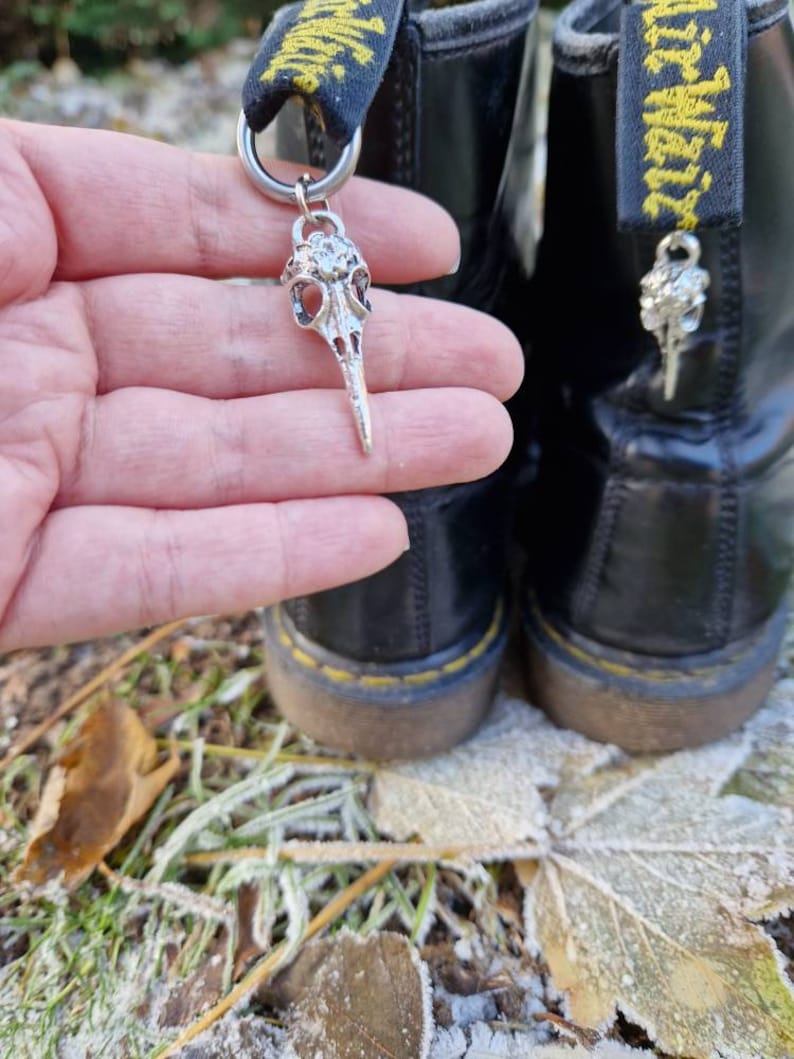 May include: A pair of black leather boots with yellow laces and a silver skull charm attached to one of the laces. The boots are sitting on a bed of fallen leaves covered in frost.