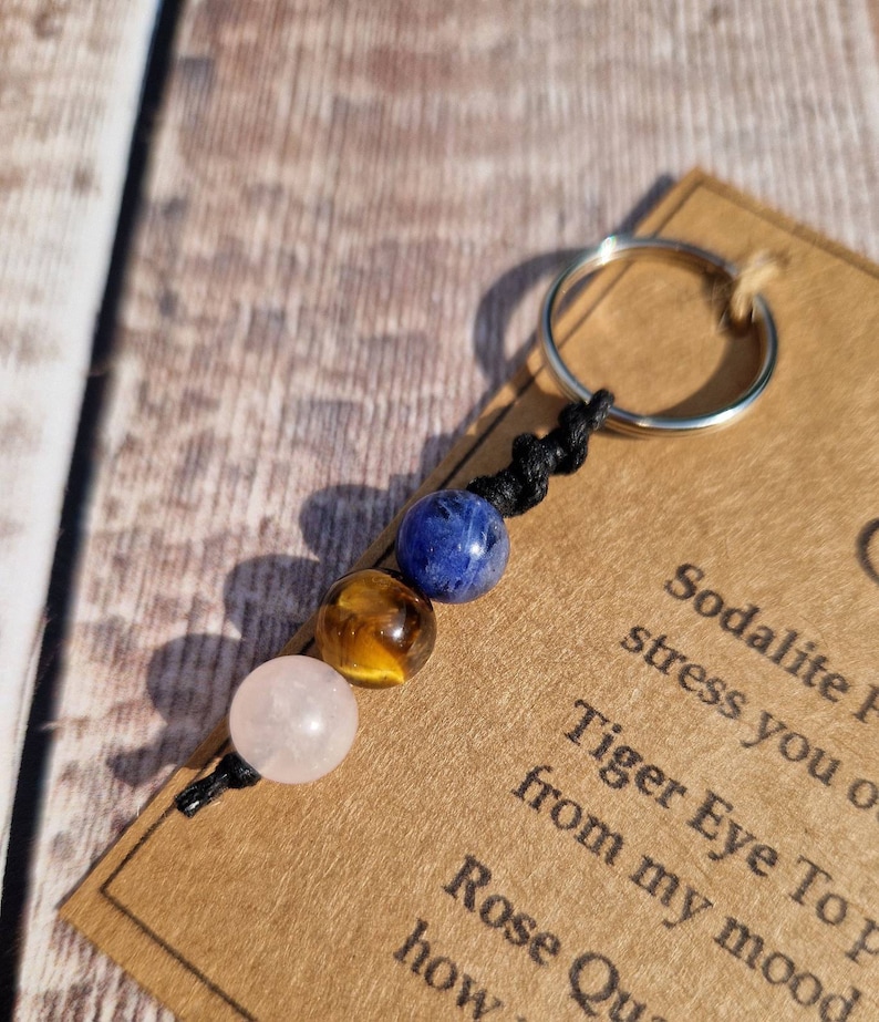 May include: A keychain with three gemstone beads: a pink rose quartz, a golden tiger's eye, and a blue sodalite. The beads are strung on black cord and attached to a silver-colored ring. A small card with text is visible.