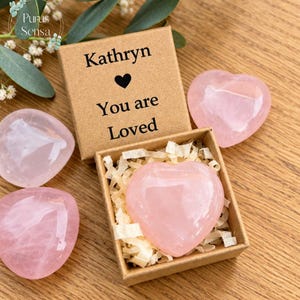 May include: A square brown cardboard gift box with the text Kathryn, a black heart icon, and the words You are Loved printed on the lid. Inside the open box sits a polished pink rose quartz heart nestled in paper filler, with two additional rose quartz hearts placed outside the box.