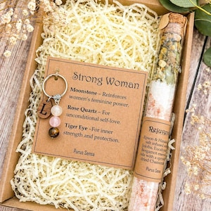 May include: A gift box containing a keychain, a test tube, and decorative filler. The keychain has a silver ring with a wooden charm and three gemstone beads. The test tube contains bath salts and dried herbs. Text on the card reads "Strong Woman".