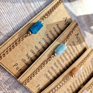 May include: Three gold chain bracelets with different colored stones. The first bracelet has a blue stone, the second has a light blue stone, and the third has a light brown stone. Each bracelet has a card attached with the name of the stone and its associated zodiac sign. The cards read "Apatite", "Aquamarine", and "Sunstone".