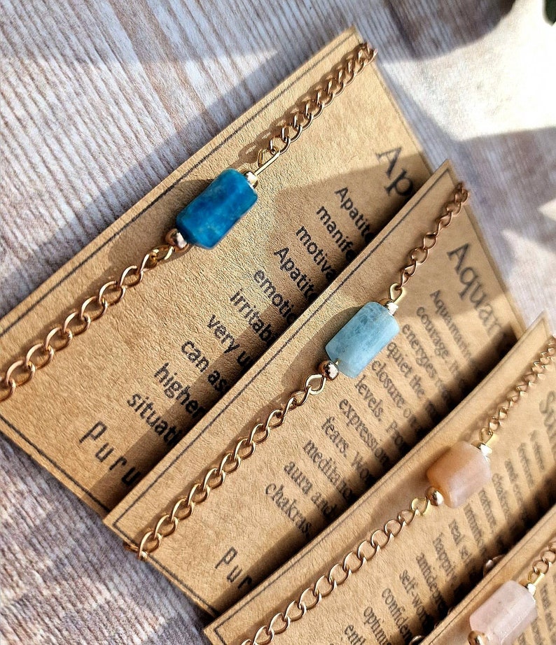May include: Three gold chain bracelets with different colored stones. The first bracelet has a blue stone, the second has a light blue stone, and the third has a light brown stone. Each bracelet has a card attached with the name of the stone and its associated zodiac sign. The cards read "Apatite", "Aquamarine", and "Sunstone".