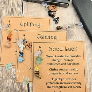 May include: A collection of gemstone keychains with informational cards. The keychains feature various colorful stones, including pink, green, yellow, and brown. The cards have text that reads "Uplifting", "Calming", and "Good Luck", with descriptions of the stones' properties.