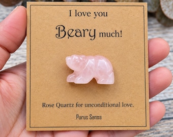 May include: A small, carved rose quartz bear figurine attached to a square tan card. The card features the black printed text: I love you Beary much! Rose Quartz for unconditional love. Purus Sensa. The bear is light pink and translucent.