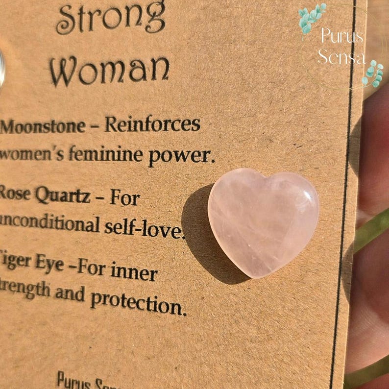 May include: A heart-shaped rose quartz gemstone on a brown card with the words "Strong Woman" and text about the properties of gemstones. The card also has the name "Purus Sensa".