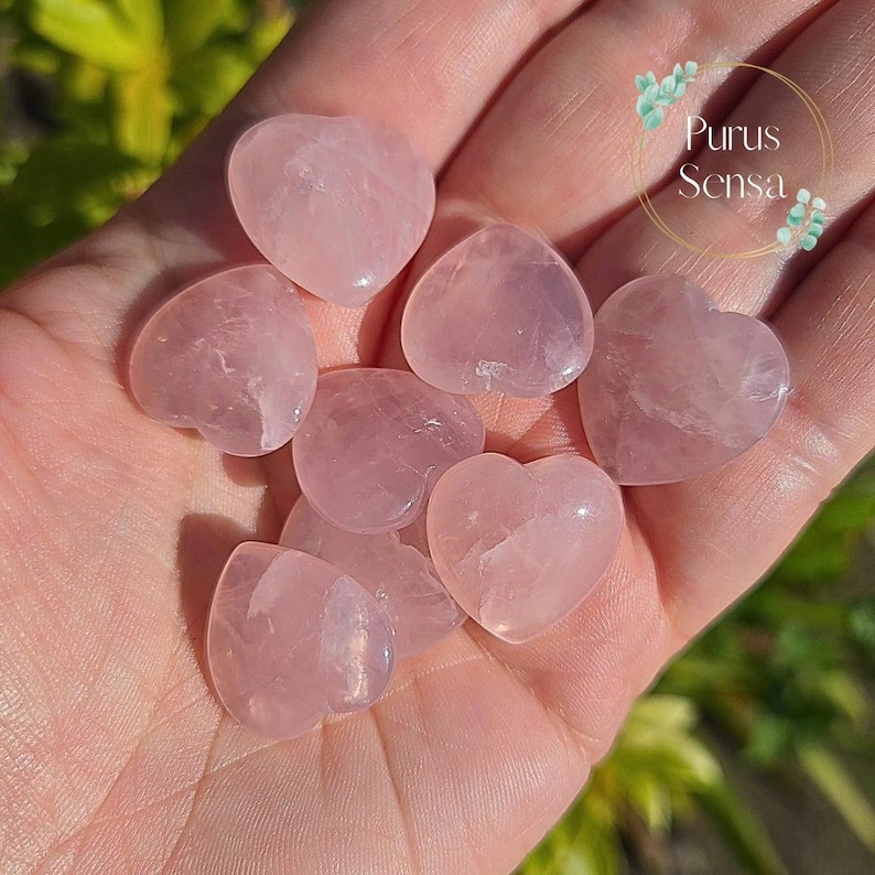 May include: A collection of polished rose quartz hearts in varying sizes. The hearts are a soft pink color and are held in an open hand. The image has a natural, sunlit appearance. The text "Purus Sensa" is in the upper right corner.