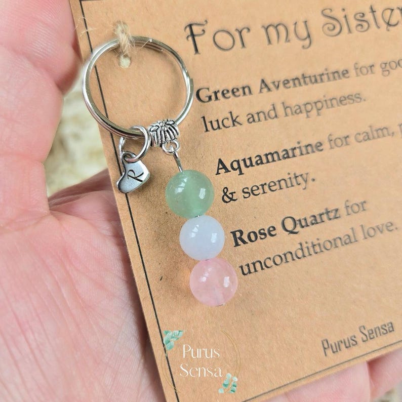 Pode incluir: Um porta-chaves com um anel prateado, um berloque de cora&ccedil;&atilde;o com a letra "R" e tr&ecirc;s contas de pedras preciosas: verde, azul claro e rosa. O cart&atilde;o diz "For my Sister".