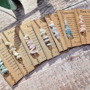 May include: A set of ten gemstone bracelets with gold chains. Each bracelet has a different type of gemstone and a card with the name of the stone and its properties. The stones include Larimar, Moonstone, Kunzite, Morganite, Blue Lace Agate, Aquamarine, and Pink Opal.