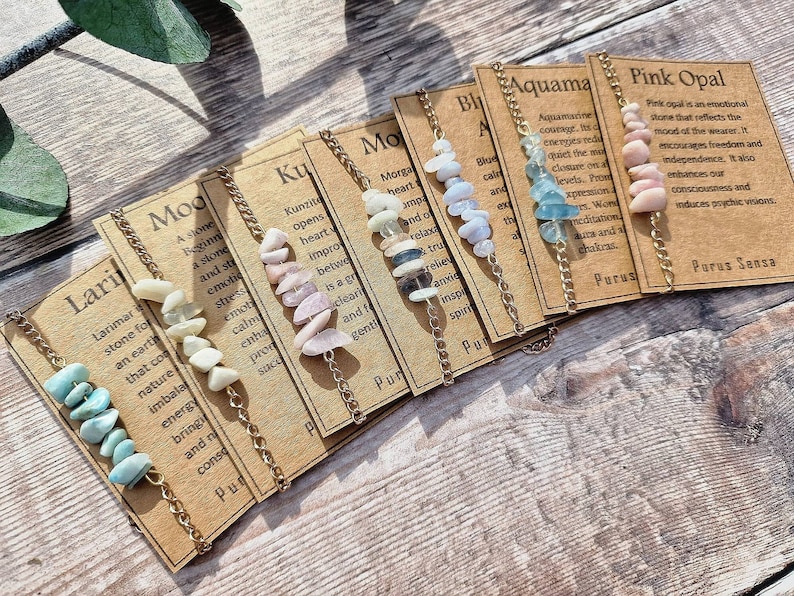 May include: A set of ten gemstone bracelets with gold chains. Each bracelet has a different type of gemstone and a card with the name of the stone and its properties. The stones include Larimar, Moonstone, Kunzite, Morganite, Blue Lace Agate, Aquamarine, and Pink Opal.