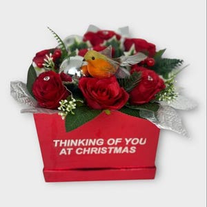 May include: A festive Christmas floral arrangement in a red box. The arrangement features red roses, silver accents, and a decorative bird. The box has the text "Thinking of You at Christmas".