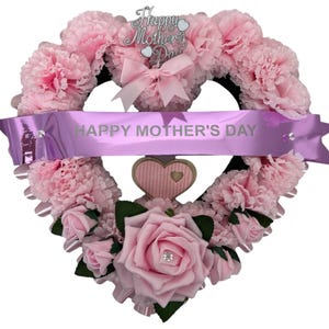 May include: A heart-shaped floral wreath with pink carnations, roses, and a lavender ribbon that reads "HAPPY MOTHER'S DAY." The wreath includes a decorative bow and a heart-shaped wooden accent.