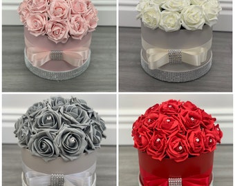 May include: Four different arrangements of artificial roses in hat boxes. The boxes are decorated with a ribbon and rhinestones. The roses are pink, white, gray, and red.