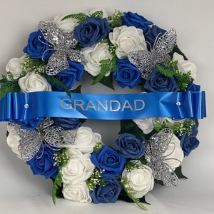 May include: A blue and white artificial flower wreath with silver butterflies and a blue ribbon with the word "GRANDAD" printed on it.
