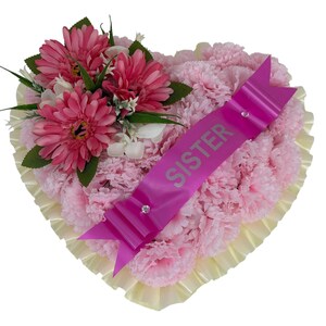 Könnte beinhalten: Ein herzförmiges Blumenarrangement aus hellrosa Nelken, einer Gruppe dunkelrosa Gerbera mit grünen Blättern und einem leuchtend pinken Schleifenband in der Mitte, auf dem das Wort SISTER in silbernen Großbuchstaben aufgedruckt ist.