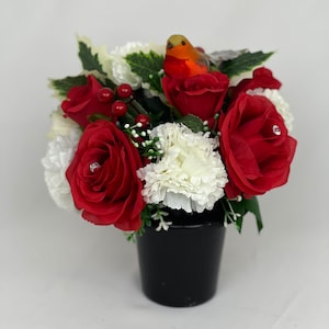 May include: A floral arrangement in a black pot. The arrangement features red roses, white carnations, and green foliage. A small, orange and brown bird figurine sits atop the flowers. The roses have a small rhinestone in the center.