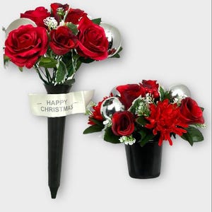 May include: Two Christmas floral arrangements. One is a tall, black vase with red roses and a banner that reads "Happy Christmas." The other is a small black pot with red flowers and silver baubles.