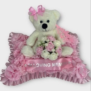 May include: A cream teddy bear with a pink bow sits on a pink floral cushion. A bouquet of pink and white roses is in front of the bear. A pink ribbon with the words "IN LOVING MEMORY" is across the front.