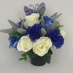 May include: A white and blue artificial flower arrangement in a black pot with a purple butterfly on top.