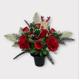 May include: A festive floral arrangement in a black pot. The arrangement features vibrant red roses, green leaves, and sprigs of holly with red berries. A wooden reindeer adds a holiday touch. The arrangement is accented with gold and silver decorative elements.