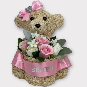 Puede incluir: Un arreglo floral rosa y blanco en un oso de sisal con un lazo rosa y un dije de corazón plateado. El oso tiene una cinta rosa con la palabra "HERMANA" impresa.