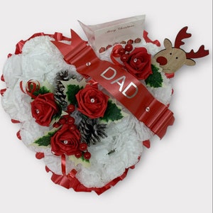Puede incluir: Arreglo floral navideño en forma de corazón con rosas rojas, bayas y una cinta roja que dice "DAD". Incluye un reno de madera y una tarjeta que dice "Merry Christmas".