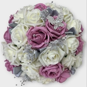 Puede incluir: Un ramo de novia hecho de rosas artificiales de marfil y rosa empolvado con detalles plateados y un broche de mariposa plateado.