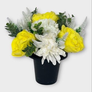May include: A yellow and white artificial flower arrangement in a black pot. The arrangement features yellow roses, white chrysanthemums, and green foliage.