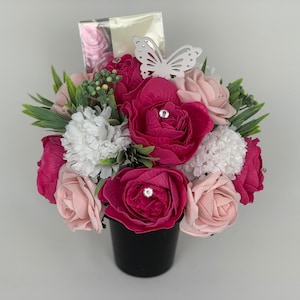 Könnte beinhalten: Ein rosa-weißer Blumenstrauß in einem schwarzen Topf mit einem dekorativen Schmetterling. Die Blumen sind aus Seife und haben Strasssteine.