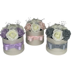 Puede incluir: Tres cajas redondas florales con lazos de cinta. Cada caja está llena de rosas de espuma en diferentes colores: morado, rosa y gris. Las cajas están decoradas con detalles plateados y un lazo de cinta.
